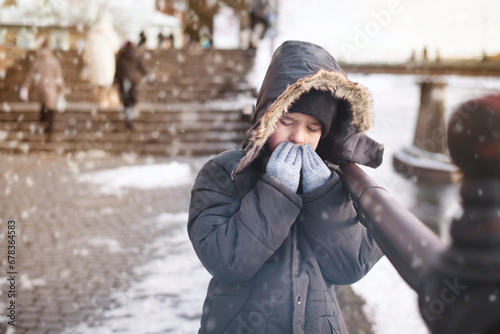 A young boy on the bank of river in winter is frozen by a strong frosty wind and blows on his hands to warm himself, although the boy is wearing gloves, but he is still cold, snow is falling photo