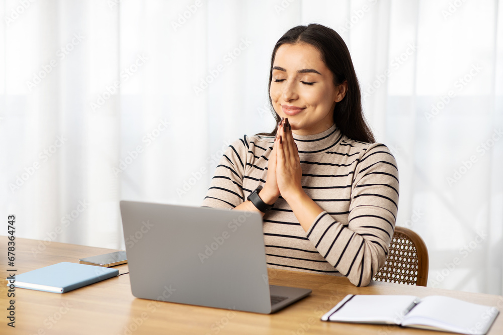 Positive young arab lady manager work at computer, make pray gesture, expect good news Positive young arab lady manager work at computer, make pray gesture, expect good news