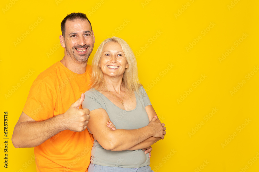 Happy adorable Caucasian couple in their 40s hugging and raising thumbs up in sign of approval and trust, isolated on yellow studio background. - obrazy, fototapety, plakaty 