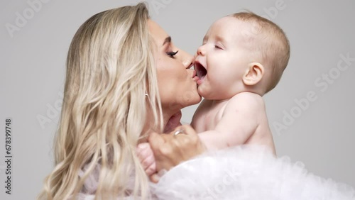 Loving mom kisses her adorable little baby. Blonde woman holds her naked infant son in studio. White backdrop. photo