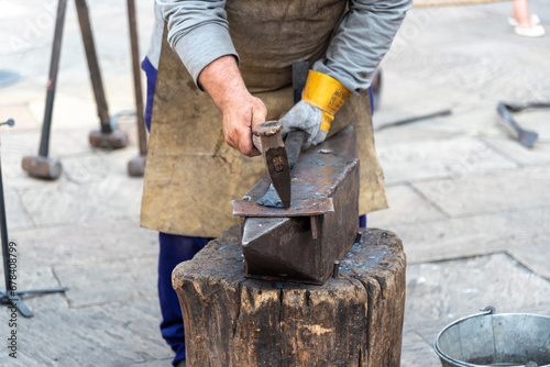 Detail of a blacksmith's hands in a brown leather apron and gloves forging a piece with a hammer on an iron anvil in the street at a street market.
