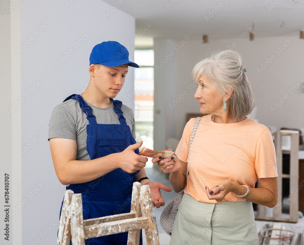 Fototapeta premium Senior woman holding bunch of cash, giving money to young man she employed for part-time work in construction site.