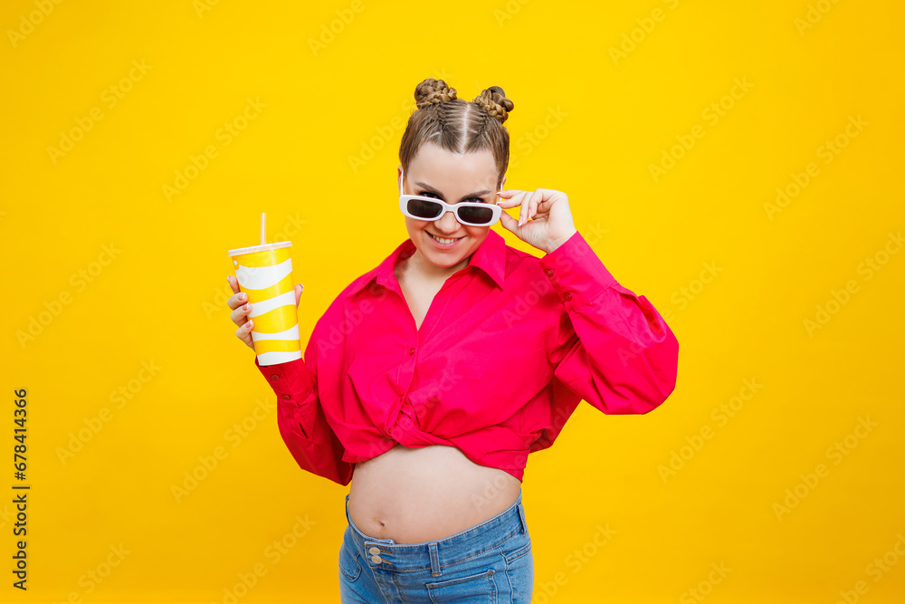 A cute pregnant woman in a pink shirt drinks water from a plastic cup. A pregnant woman on a yellow background with a glass in her hands. Harmful water for pregnant women.