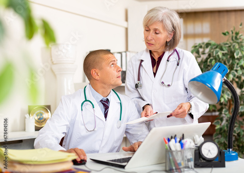 Male physician sitting at table in his office. His grey haired female coworker with medical documentation standing next to him.