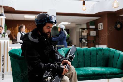 Male skier wearing snow gear sits on sofa in lounge area of winter resort, holding wintersports equipment. With skiing skis in hand, guest is ready to embark on his winter holiday adventure.