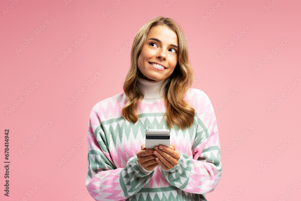 Portrait of smiling beautiful woman wearing cozy sweater, holding mobile phone looking away standing isolated on pink background. Technology, online shopping concept Portrait of smiling beautiful woman wearing cozy sweater, holding mobile phone looking away standing isolated on pink background. Technology, online shopping concept