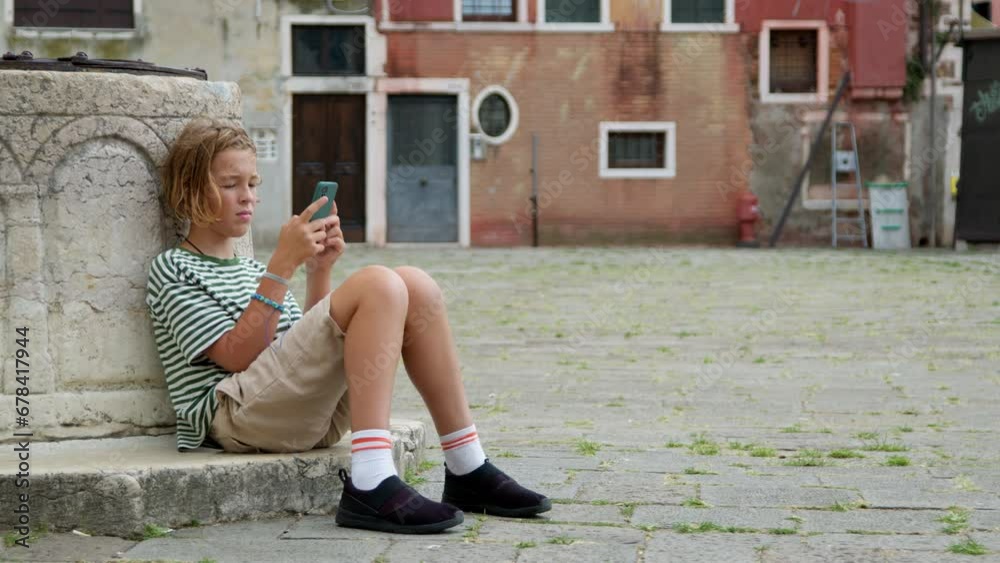 A teenager sits at a Venetian square, deeply engrossed in his smartphone. Dressed in a striped shirt and casual shorts, he seems oblivious to the historic beauty around him A teenager sits at a Venetian square, deeply engrossed in his smartphone. Dressed in a striped shirt and casual shorts, he seems oblivious to the historic beauty around him