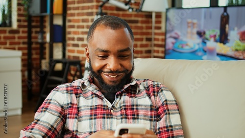Cozy african american man chatting with online friends over the phone in warm stylish home, enjoying himself. Happy smiling person scrolling on phone entertained by social media activity