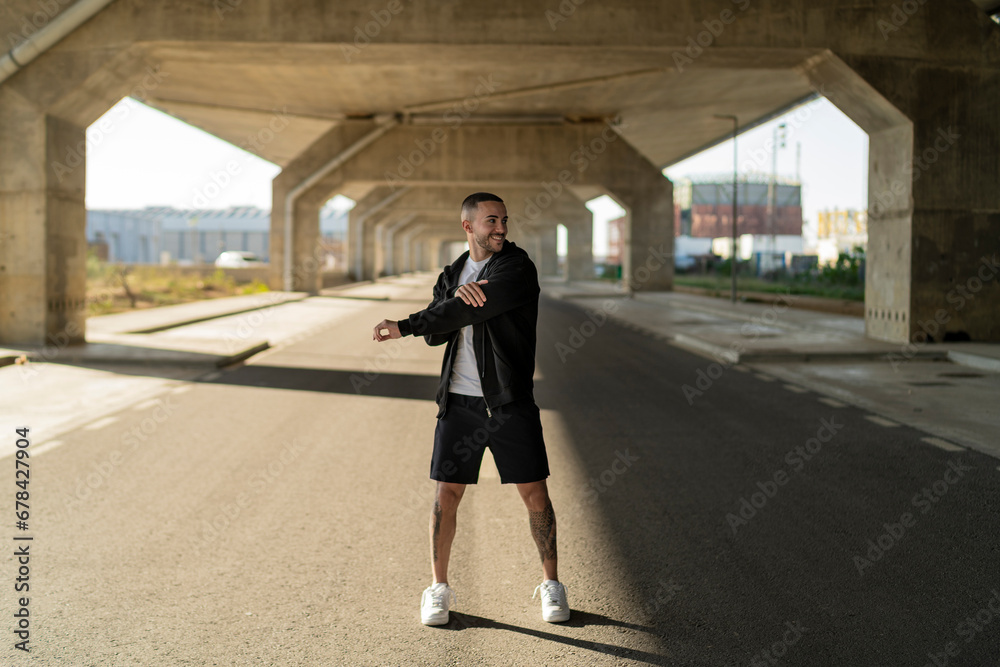 Chico joven tatuado y musculoso posando y haciendo deporte en la calle en un tunel con un puente de fondo