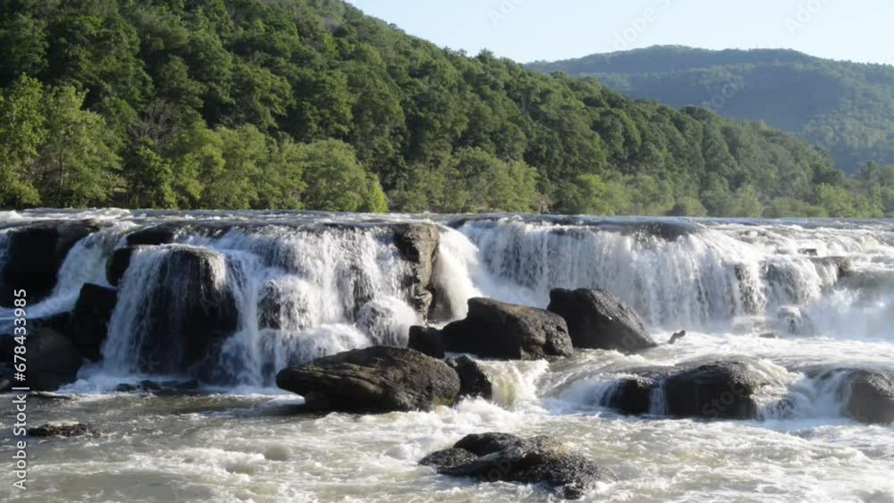 waterfall spanning across river with boulders, summer trees, and a mountain range in the background. waterfall spanning across river with boulders, summer trees, and a mountain range in the background.