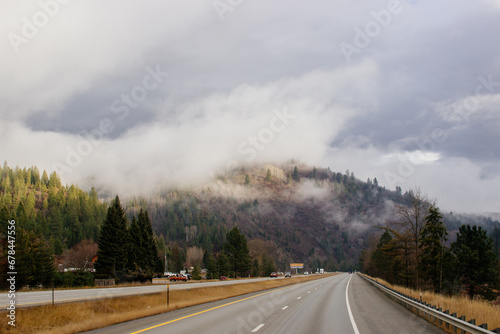 Asphalt road in the middle of high mountains, covered with fog and clouds on autumn day. American winter landscape of a mountainous area covered with fir forest. Fall season on the highway with cars © Liudmila