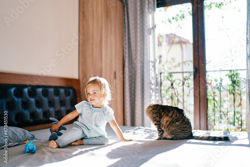 Little girl sits on a bed with a toy in her hand near a tabby cat