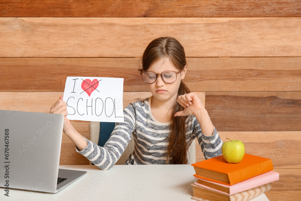 Upset little girl holding paper with text I DO NOT LOVE SCHOOL at table on wooden background Upset little girl holding paper with text I DO NOT LOVE SCHOOL at table on wooden background