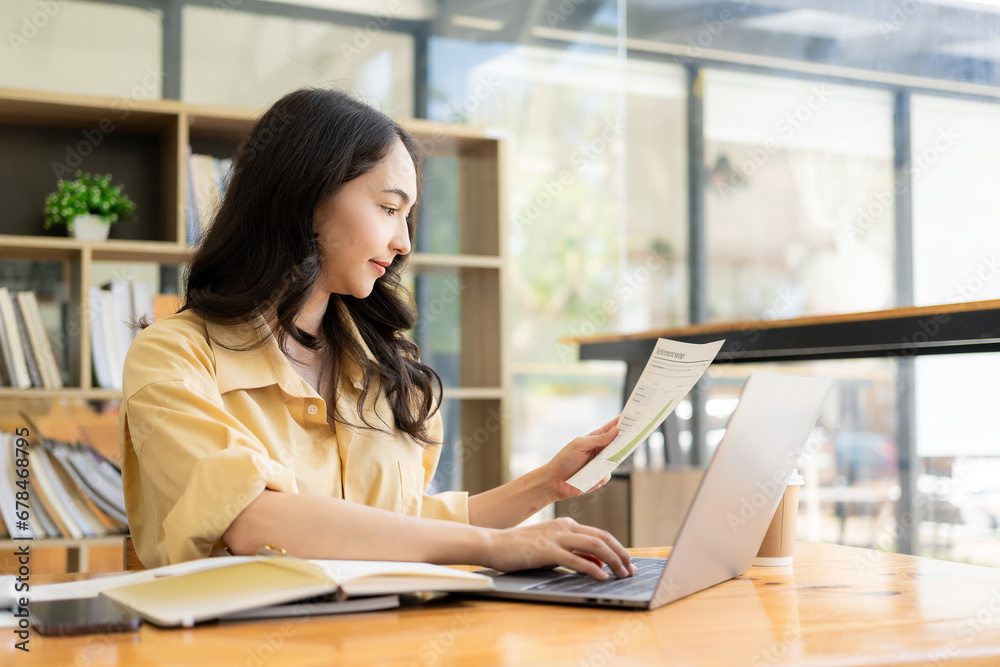 © ArLawKa - Ambitious Asian girl working from home Looking at laptop screen and financial documents, woman is checking email or doing research while working remotely.