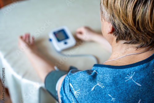 Senior woman checking blood pressure level at home, older female suffering from high blood pressure using a pulsometer, tonometer. Back view photo