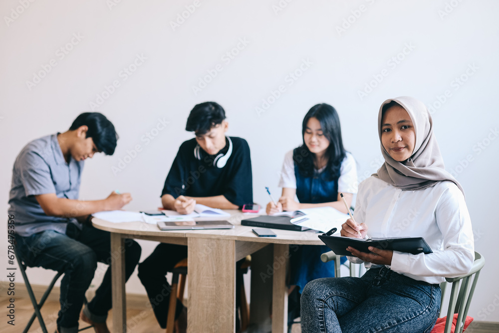 Muslim Asian student girl holding a book and smiling at the camera while classmates are studying in the background Muslim Asian student girl holding a book and smiling at the camera while classmates are studying in the background