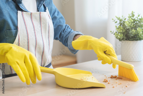 Household clean up, housekeeper asian young woman wearing protection rubber yellow gloves, using broom, dustpan sweeping remove spilled crumb food broken on dirty table at home, equipment for cleaning photo