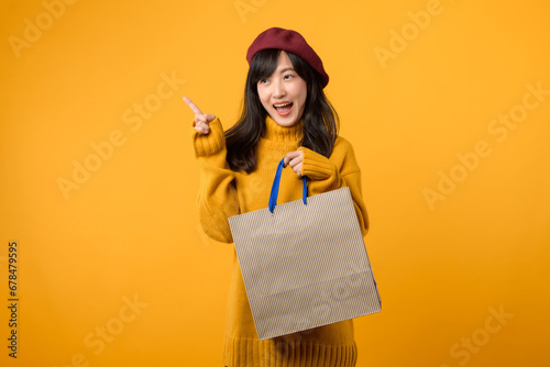 Young Asian woman in her 30s, wearing a yellow sweater and red beret, indulges in a shopping extravaganza and pointing finger to free copy space against a vibrant yellow backdrop. photo