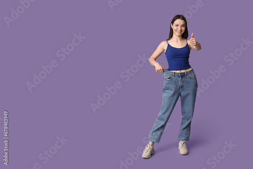 Beautiful young sporty woman in loose jeans showing thumb-up gesture on purple background. Weight loss concept