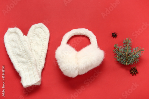 Warm mittens, earmuffs and fir branch on red background