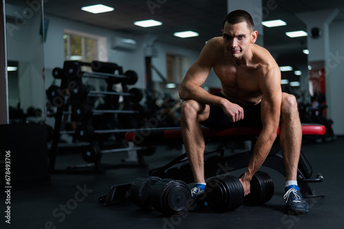 Muscular shirtless man sitting on a bench and lifting a dumbbell. 