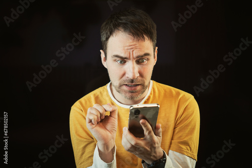 Bad news in smartphone. man looks at a message on his mobile phone with wide eyes and an open mouth. He is shocked and scared. Human emotions on black background