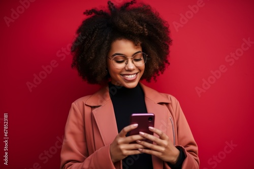 Studio portrait of beautiful African American woman with smartphone in pink clothes against red background. Positive girl with Afro haircut texting message, enjoying online communication, using app.
