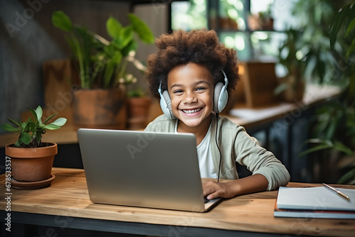 Smiling African American boy sitting at desk with laptop and headphones in cozy room. Smart black kid studying online lesson, participating in web meeting with tutor. Home schooling concept.