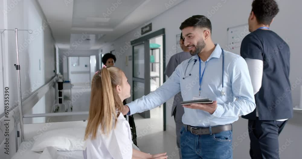 Biracial male doctor and caucasian girl patient sitting on bed talking in hospital, slow motion Biracial male doctor and caucasian girl patient sitting on bed talking in hospital, slow motion