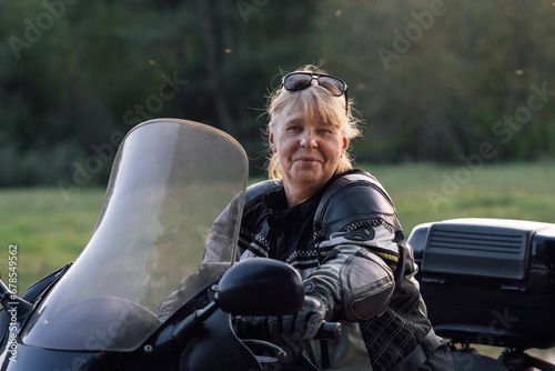 Happy smiling blonde middle-aged motorcyclist in leather jacket,gloves is sitting on her bike .