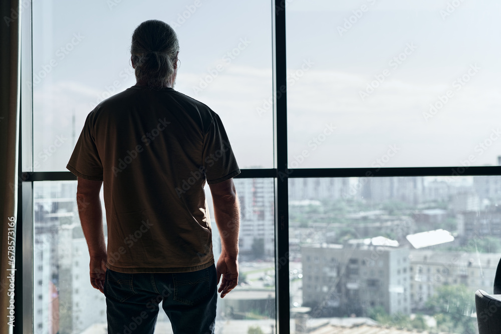 Rear view of mature man standing in front of panoramic window and looking at the city Rear view of mature man standing in front of panoramic window and looking at the city