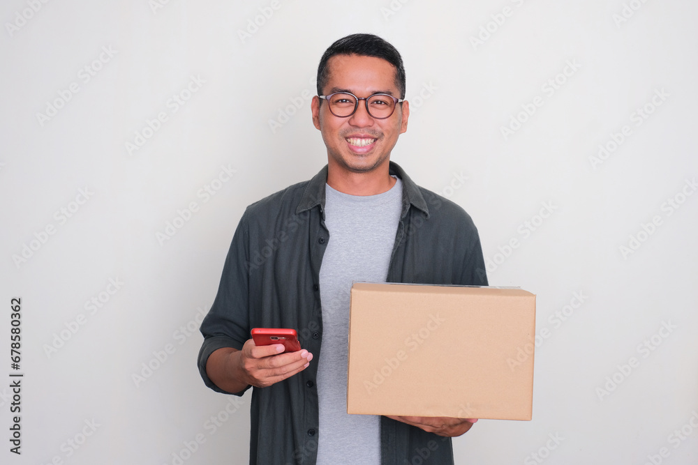 Adult Asian man smiling while holding a package box and mobile phone Adult Asian man smiling while holding a package box and mobile phone