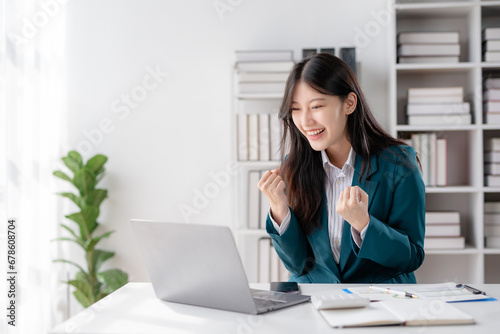 Excited cheerful asian business woman using laptop computer in the office, getting good news, feeling joy, laughing, making winner gesture, happy to win prize. © amnaj
