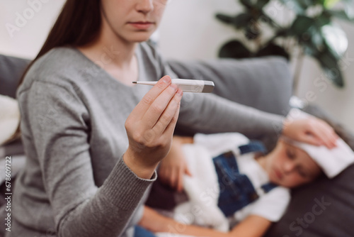 Selective focus of young mother treating sick daughter kid suffering from flu and fever, holding thermometer, checking high body temperature, applying cold compress on forehead. Childcare concept