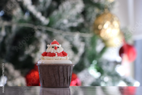 Cupcakes decorated with buttercream. It is a Santa Claus pattern in a brown cup. Placed on out focus background of Christmas trees with white snow, Golden and red balls.