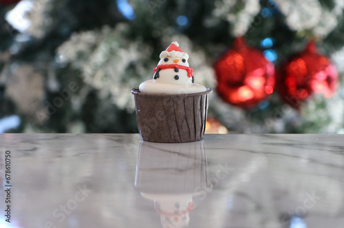 Cupcakes decorated with buttercream. It is a Snowman pattern in a brown cup. Placed on out focus background of Christmas trees with white snow and two red balls.