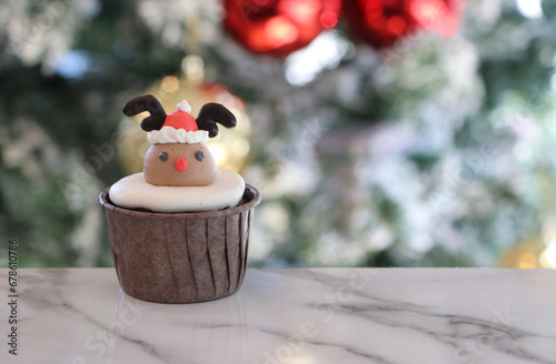 Cupcakes decorated with buttercream. It is a reindeer pattern and a Christmas hat on the head, in a brown cup. Placed on out focus background of Christmas trees with white snow, golden and red balls.