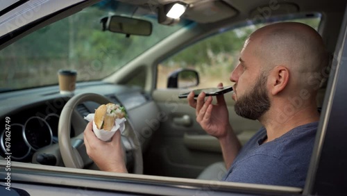 Attractive man using mobile phone, talking on the speaker and eating burger while driving car. Woman talking on in-car speakerphone while driving photo
