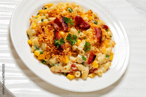 close-up of portion of macaroni and cheese with corn, bacon topped with panko breadcrumbs on white plate on white wooden table, dutch angle view