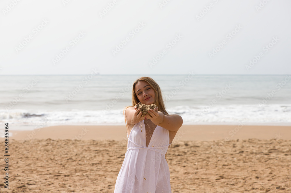Pretty young blonde woman in white dress spills sand from between her hands as she holds her hands out in front of her. The woman is happy and having fun. In the background the blue sea. Pretty young blonde woman in white dress spills sand from between her hands as she holds her hands out in front of her. The woman is happy and having fun. In the background the blue sea.