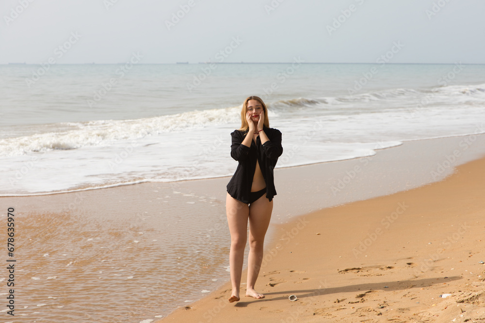 Young and beautiful blonde woman walks along the shore of the beach dressed in black shirt and black bikini and makes different expressions and postures. In the background the sea and the sky. Young and beautiful blonde woman walks along the shore of the beach dressed in black shirt and black bikini and makes different expressions and postures. In the background the sea and the sky.