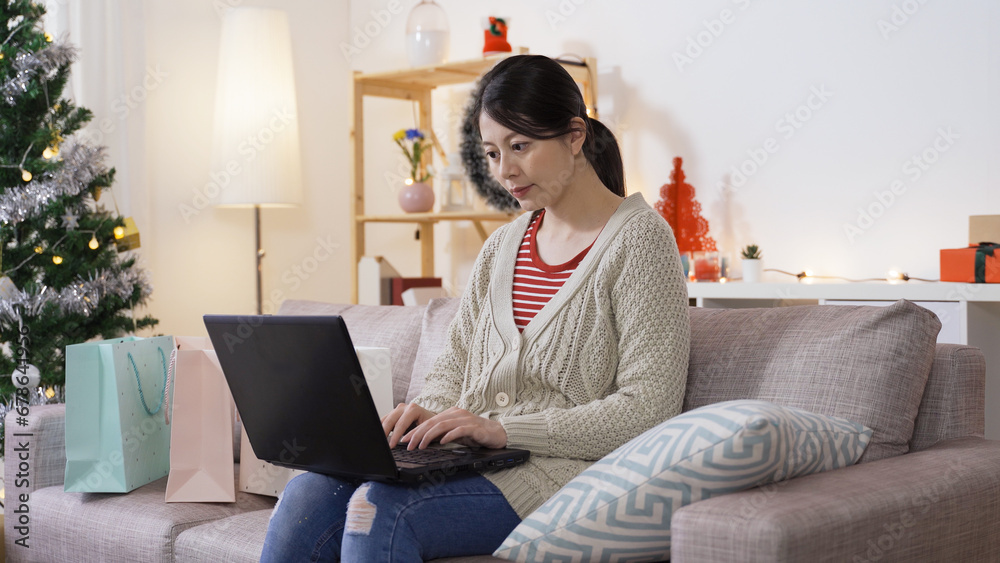 cheerful asian lady sitting in living room with festive decoration is making a shopping list for Christmas on laptop in the morning. cheerful asian lady sitting in living room with festive decoration is making a shopping list for Christmas on laptop in the morning.