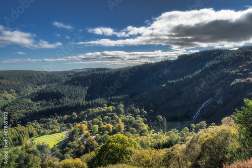 Tall Powerscourt Waterfall, valley, forests in autumn colours illuminated by sunlight. Scenic Wicklow Mountains, Ireland