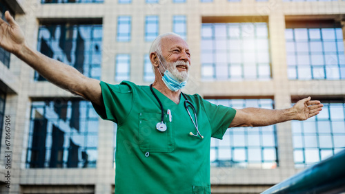 A mature doctor is standing in front of the hospital with open arms and feeling relieved after a hard day at work.