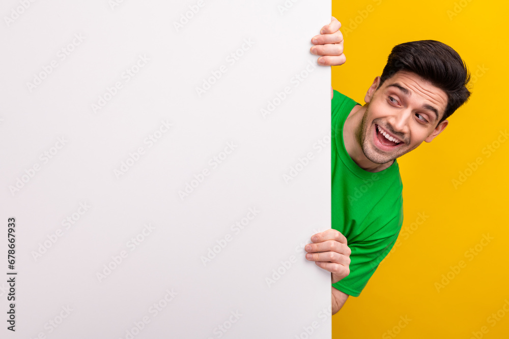 Portrait of good mood overjoyed guy wear stylish t-shirt look at offer on white billboard empty space isolated on yellow color background Portrait of good mood overjoyed guy wear stylish t-shirt look at offer on white billboard empty space isolated on yellow color background