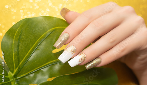 Female hand with glitter nail polish. Nail design in gold and white shades. Women hand with sparkle manicure and green leaf