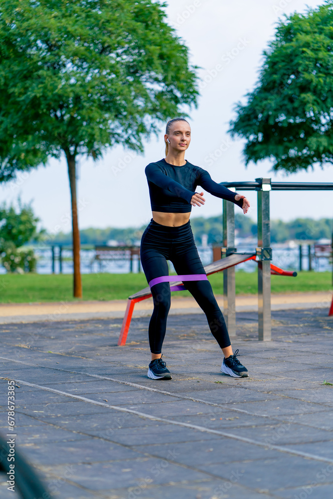 Beautiful girl in sports uniform performs exercises to pump up her legs with rubber band on a sports street playground