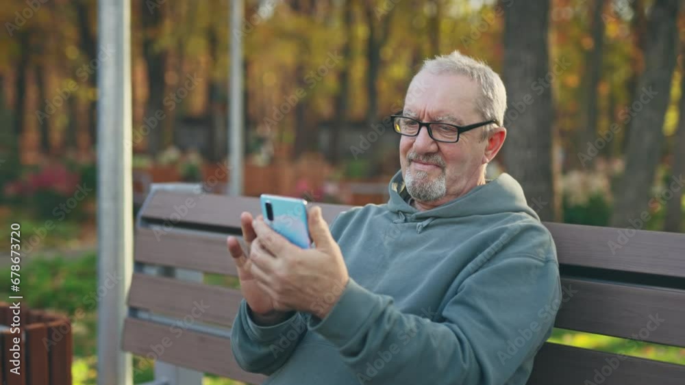 An elderly man uses a smartphone while sitting on a park bench. Pensioner surfing the Internet with a smartphone An elderly man uses a smartphone while sitting on a park bench. Pensioner surfing the Internet with a smartphone
