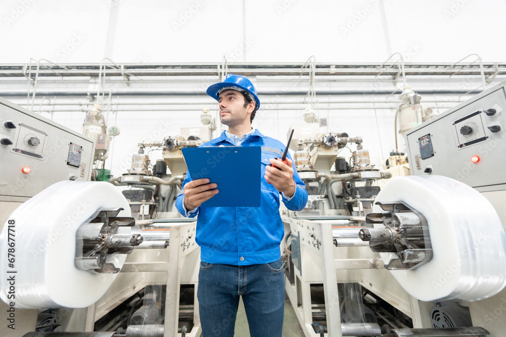 American male engineer, manager, technician Standing close and inspecting the plastic and steel production machinery of an industrial factory, holding a notepad, radio, wearing a hard hat and uniform.