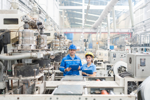 American male engineer and Asian female manager Wear a uniform and safety helmet. Standing in the middle of several working machines producing plastic and steel. In industrial business factories